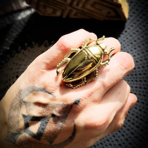 Op de afbeelding: Een gouden keverring met ingewikkelde details. De ring wordt gedragen op een hand met een tatoeage.