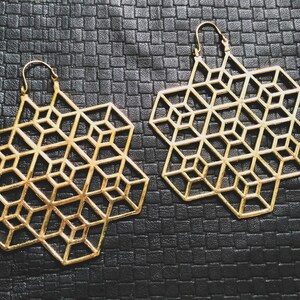Puede incluir: Un par de pendientes geométricos dorados con un diseño de cubo 3D. Los pendientes son grandes y cuelgan de la oreja.