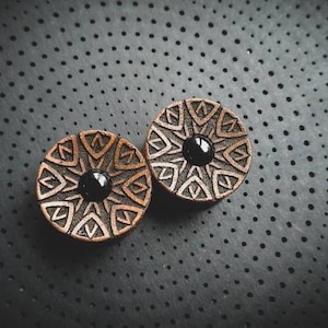 Può includere: Due tappi per orecchie in legno con intarsi in pietra nera e un motivo geometrico intagliato.