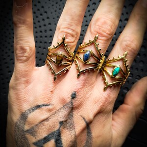 Könnte beinhalten: Drei goldene Ringe mit verschiedenen Edelsteinen. Die Ringe sind mit einem geometrischen Muster verziert und haben eine spitze Oberseite. Die Edelsteine sind ein Tigerauge, ein blauer Stein und ein türkisfarbener Stein.