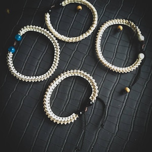 Peut inclure: Quatre bracelets en os blanc avec des liens en ficelle noire et de petites perles. Les bracelets sont sur une surface textur&eacute;e noire.