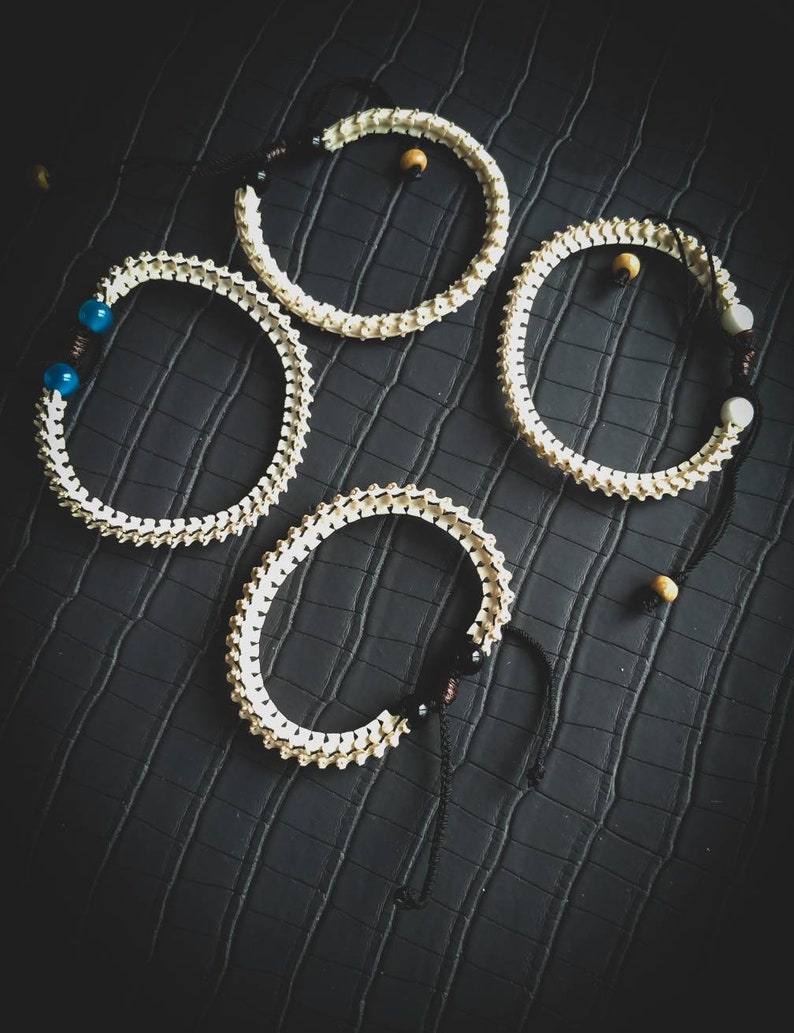Peut inclure: Quatre bracelets en os blanc avec des liens en ficelle noire et de petites perles. Les bracelets sont sur une surface textur&eacute;e noire.