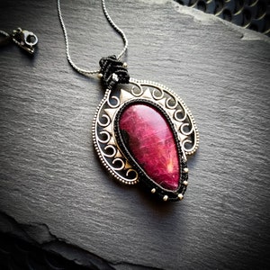 Peut inclure: Un collier avec un pendentif en argent orné d'une pierre précieuse rouge foncé dans un motif en filigrane argenté. Le collier est sur un fond noir.