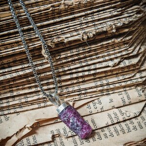 Peut inclure: Un collier en argent avec un pendentif cylindrique violet foncé. Le pendentif est fait d'un matériau rugueux et texturé. Le collier repose sur les pages d'un vieux livre ouvert avec du papier vieilli et du texte.