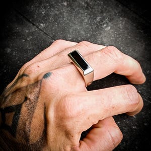 Peut inclure: Une bague en argent avec une pierre d'onyx noire rectangulaire. La bague a un design moderne et minimaliste. L'anneau est épais et poli, et la pierre noire est au ras de la surface. Cette bague est une pièce de caractère.