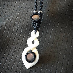 以下が含まれることがあります： 2つの濃いグレーの宝石ビーズでアクセントが付けられた、ダブルインフィニティシンボルデザインの白い骨のペンダント。ペンダントは黒いコードネックレスに取り付けられています。