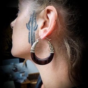 Peut inclure: Une paire de grandes boucles d'oreilles en bois en forme de cercle avec des accents en métal doré. Les boucles d'oreilles sont brunes et ont une finition lisse et polie.