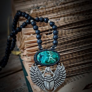 May include: A black lava rock beaded necklace with a silver scarab beetle pendant. The pendant features a green stone with a marbled pattern.