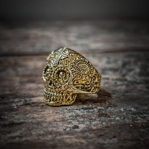 Peut inclure: Une bague en forme de crâne en or avec des détails complexes et un motif floral. La bague est en métal et a une finition polie.