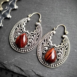 Peut inclure: Une paire de boucles d'oreilles créoles en argent avec des motifs floraux complexes et des pierres de œil de tigre rouge.