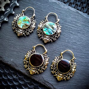 Peut inclure: Quatre paires de boucles d'oreilles créoles en or avec un design circulaire représentant plusieurs visages et une pierre centrale. Deux paires ont une pierre en nacre d'ormeau verte, et deux paires ont une pierre noire.