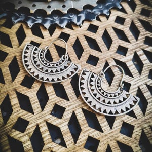 May include: A pair of silver hoop earrings with a geometric design. The earrings are on a wooden surface with a diamond pattern.