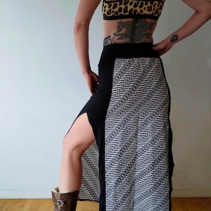 Peut inclure: Une jupe maxi noire avec une fente sur le côté et un motif géométrique blanc et noir sur le côté droit. La jupe est portée avec des bottes brunes.