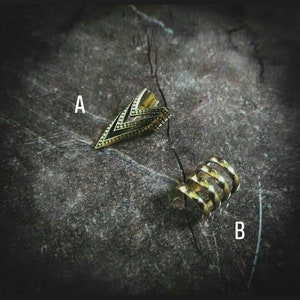 May include: Two gold ear cuffs, one with a geometric triangle design and the other with a spiral design. The cuffs are on a dark grey textured surface.