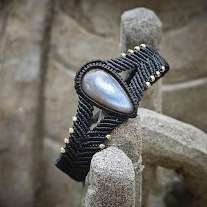 Puede incluir: Un brazalete de macramé negro con una piedra preciosa gris plateada. El brazalete está adornado con pequeñas cuentas plateadas.