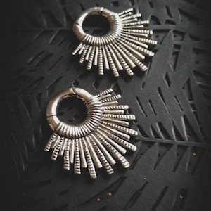 May include: Two silver ear gauges with a sunburst design. The gauges are on a black background with a geometric pattern.