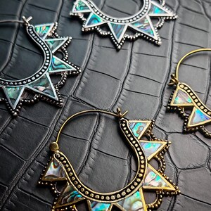 Peut inclure: Gros plan sur quatre boucles d'oreilles créoles avec un motif de soleil. Les boucles d'oreilles présentent un cadre en métal argenté ou doré avec des accents triangulaires et des incrustations de nacre irisée. Le fond est une surface noire texturée.