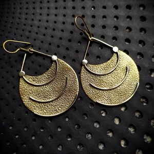 May include: A pair of gold-toned earrings with a textured finish. The earrings are shaped like a crescent moon with three horizontal lines. The earrings are hanging from a black background.