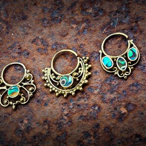 Op de afbeelding: Drie sierlijke, goudkleurige septumringen met iriserende abalone schelpinleg. De ringen hebben ingewikkelde filigraan en decoratieve elementen. De sieraden worden getoond op een getextureerd, roestbruin oppervlak.