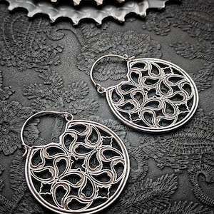 Peut inclure: Une paire de boucles d'oreilles ajourées argentées avec un motif circulaire présentant un motif tourbillonnant complexe. Les boucles d'oreilles sont posées sur un fond noir texturé, avec un pignon de vélo visible en haut.