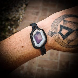 Peut inclure: Un bracelet en macramé noir orné d'une grande pierre d'améthyste rectangulaire avec une bordure blanche et un centre violet. Le bracelet est porté sur un poignet avec un tatouage.