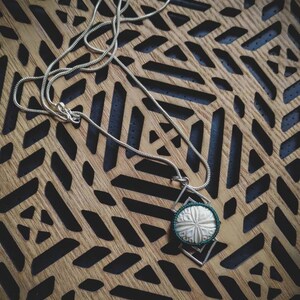 Puede incluir: Un collar de cadena de plata con un colgante que presenta un círculo blanco tallado con un borde verde, engastado en un marco de plata.
