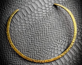 Puede incluir: Un collar de metal dorado martillado. El diseño circular abierto se asienta sobre un fondo texturizado gris oscuro. El collar tiene un diseño simple y elegante.