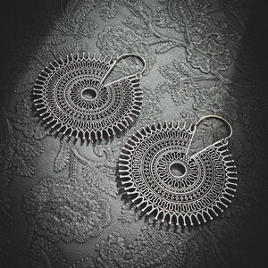 Peut inclure: Deux boucles d'oreilles circulaires argentées avec des motifs symétriques complexes. Chaque boucle d'oreille présente un cercle central entouré de motifs détaillés et un crochet. Les boucles d'oreilles sont posées sur une surface sombre et texturée.