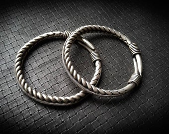 May include: Two silver braided bracelets with a twisted design. The bracelets are on a black textured surface.