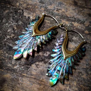 Può includere: Un paio di grandi orecchini a cerchio con una cornice in metallo dorato e ali di madreperla di abalone iridescenti. Gli orecchini sono su una superficie di legno.