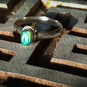 Peut inclure: Une bague en argent ornée d'une pierre précieuse ovale et irisée. La bague est posée sur une surface en bois sombre sculptée avec un motif de labyrinthe. La pierre affiche des nuances de vert et de bleu.