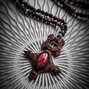 May include: A wooden pendant with a red gemstone in the center, hanging from a black and brown beaded necklace. The pendant is carved with intricate details and has a circular opening at the top.