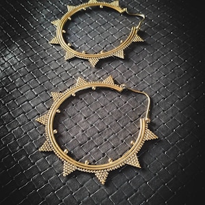 Peut inclure: Deux boucles d'oreilles dorées en forme de créoles, ornées d'un motif de soleil. Les boucles d'oreilles présentent de petites perles rondes et des pointes triangulaires. Elles sont posées sur un fond sombre et texturé.