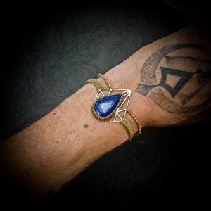 Puede incluir: Una pulsera dorada con una piedra preciosa azul en forma de lágrima, engastada en un marco geométrico. La pulsera tiene una banda tejida y se lleva en la muñeca. El fondo es oscuro.