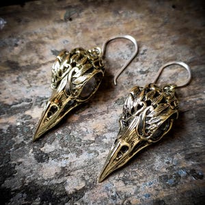 Peut inclure: Une paire de boucles d'oreilles en or ton sur ton en forme de crânes d'oiseaux avec des détails complexes. Les boucles d'oreilles ont une fermeture à crochet argentée.