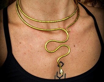 May include: A gold-colored necklace in the shape of a snake. The necklace has two bands around the neck and a snake design that dangles down the chest. The necklace is worn over a black tank top.