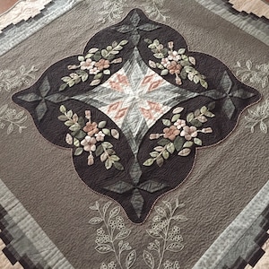 Può includere: Una coperta trapuntata dettagliata con un motivo floreale centrale. La trapunta presenta un motivo a forma di diamante al centro, circondato da un bordo marrone scuro con abbellimenti floreali. La tavolozza dei colori include sfumature di marrone, grigio e crema.