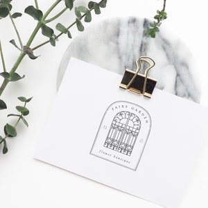 May include: A white card with a black and white logo for a flower boutique called "Fairy Garden". The logo features a decorative archway with a gate. The card is held by a gold binder clip on a marble surface.