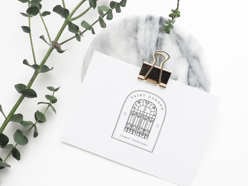 May include: A white card with a black and white logo for a flower boutique called "Fairy Garden". The logo features a decorative archway with a gate. The card is held by a gold binder clip on a marble surface.