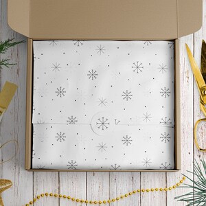 May include: A brown cardboard box with a white snowflake patterned wrapping paper inside. The box is open and the wrapping paper is visible. There is a gold pair of scissors and a gold string of beads on a white wooden surface.