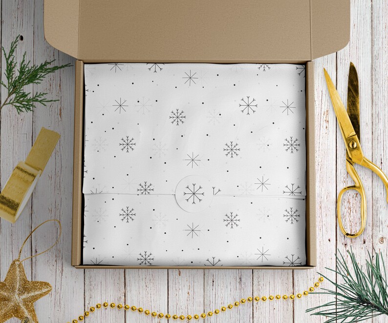 May include: A brown cardboard box with a white snowflake patterned wrapping paper inside. The box is open and the wrapping paper is visible. There is a gold pair of scissors and a gold string of beads on a white wooden surface.