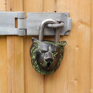 Puede incluir: Un candado decorativo con dise&ntilde;o de cabeza de oso, en bronce y verde, est&aacute; asegurado a una puerta de madera con un pestillo de metal. El grillete en forma de U del candado es plateado, y la puerta est&aacute; hecha de tablones de madera verticales.