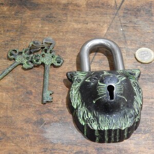 Puede incluir: Un candado decorativo negro con un dise&ntilde;o de cabeza de le&oacute;n, acompa&ntilde;ado de dos llaves ornamentadas. El candado tiene un grillete plateado. Las llaves y el candado tienen una p&aacute;tina verde. Una moneda est&aacute; al lado del candado.