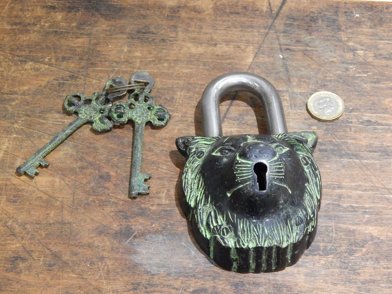 Puede incluir: Un candado decorativo negro con un dise&ntilde;o de cabeza de le&oacute;n, acompa&ntilde;ado de dos llaves ornamentadas. El candado tiene un grillete plateado. Las llaves y el candado tienen una p&aacute;tina verde. Una moneda est&aacute; al lado del candado.