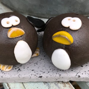 Puede incluir: Dos figuras de pingüinos de cerámica con cuerpos marrones, vientres blancos y picos amarillos. Los pingüinos tienen ojos blancos y alas negras. Están sobre una base gris texturizada, ideal para la decoración del hogar.