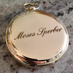 May include: A gold-coloured pocket watch with the name "Moses Sperber" engraved on the cover. The watch has a circular shape with a small loop at the top. The watch is on a speckled grey surface.
