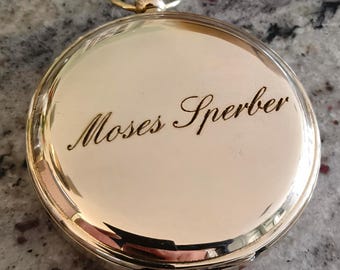 May include: A gold-colored pocket watch with the name "Moses Sperber" engraved on the cover. The watch has a circular shape with a small loop at the top. The watch is on a speckled gray surface.