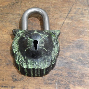 Puede incluir: Un candado negro con un dise&ntilde;o de cabeza de le&oacute;n y un grillete plateado. Los rasgos del le&oacute;n se destacan con acentos verdes. El candado est&aacute; sobre una superficie de madera.