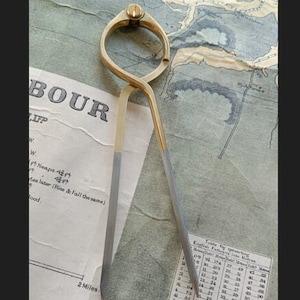 以下が含まれることがあります： ヴィンテージの真鍮とスチールのディバイダー（航海用具）が、古い地図の上に置かれています。ディバイダーは、ネジ調整付きの湾曲した上部と、2つの尖った脚を持っています。地図には「BOUR」や「2 Miles」などの文字が含まれています。