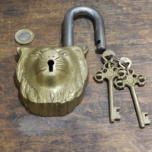 Puede incluir: Un candado de lat&oacute;n vintage con un dise&ntilde;o de cabeza de le&oacute;n y un grillete de acero abierto. Dos llaves ornamentadas est&aacute;n al lado del candado. El candado est&aacute; sobre una superficie de madera.